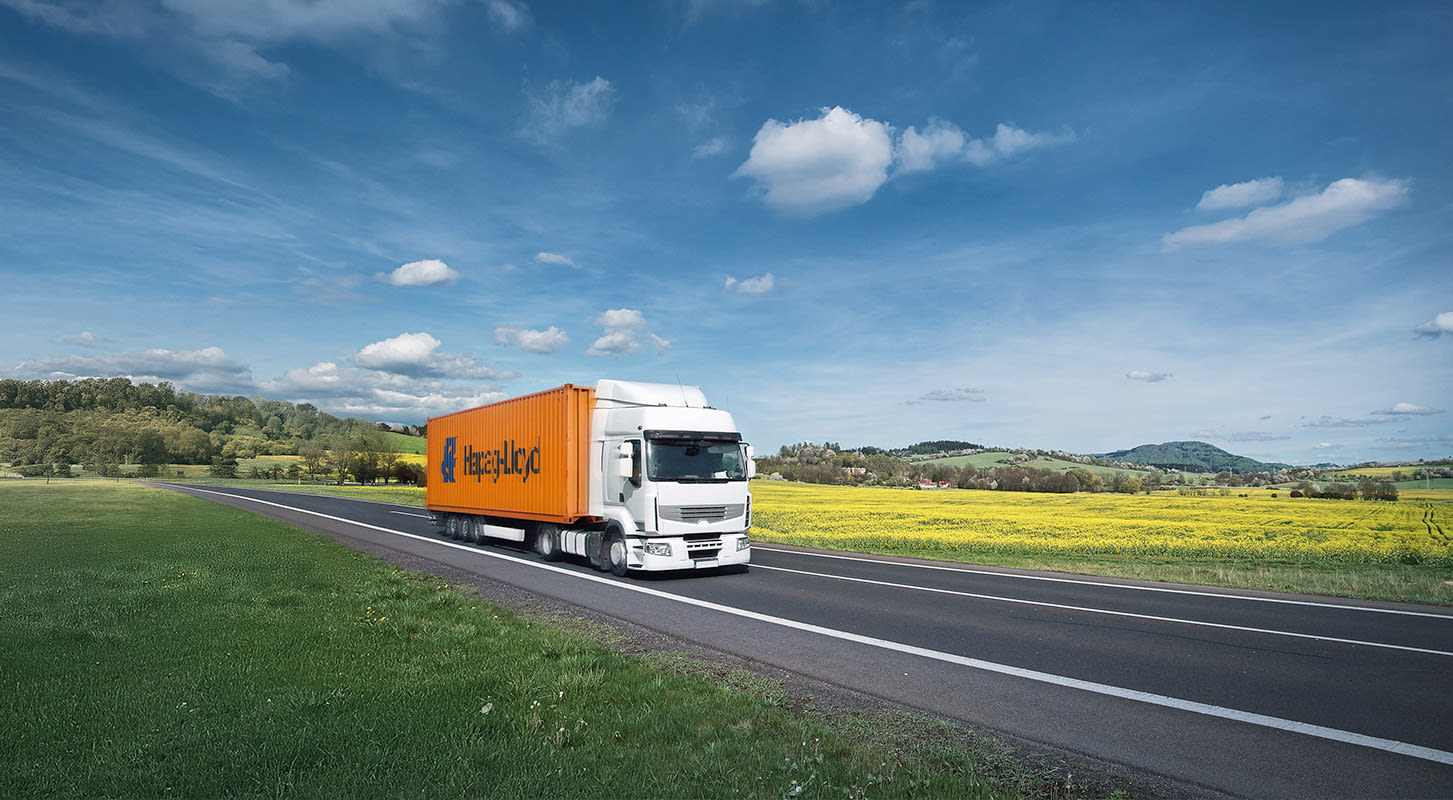 A Hapag-Lloyd orange truck is seen driving on a road beside a vibrant green field.
